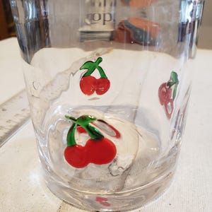 May include: A clear glass tumbler with red cherry designs and green stems. The cherries are painted on the outside of the glass. The tumbler is cylindrical and has a thick base.