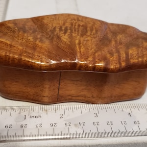 May include: A small, wooden box with a unique, wavy, and irregular shape. The box is made of light brown wood with darker brown streaks and a glossy finish. The lid is slightly raised and has a similar wavy shape to the base.