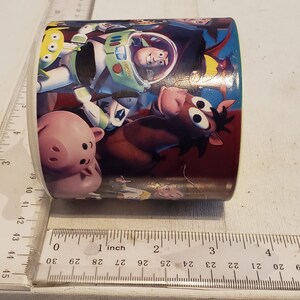 May include: A white ceramic mug with a colorful design featuring characters from the Toy Story movie franchise. The design includes Woody, Buzz Lightyear, Hamm, and Bullseye. The mug is approximately 3 inches in diameter.