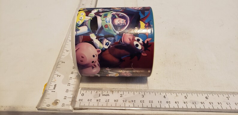 May include: A white ceramic mug with a colorful design featuring characters from the Toy Story movie franchise. The design includes Woody, Buzz Lightyear, Hamm, and Bullseye. The mug is approximately 3 inches in diameter.
