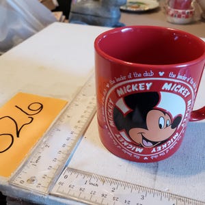 Puede incluir: Una taza de cer&aacute;mica roja con Mickey Mouse. La taza tiene un &oacute;valo blanco con la cara de Mickey y la palabra "MICKEY" repetida alrededor del &oacute;valo. El texto "the leader of the club" tambi&eacute;n est&aacute; impreso en la taza.