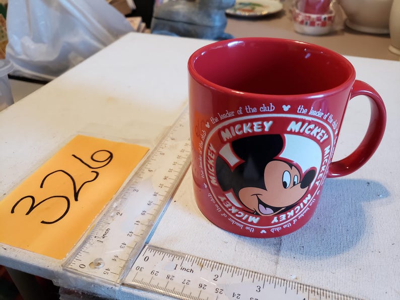 Puede incluir: Una taza de cer&aacute;mica roja con Mickey Mouse. La taza tiene un &oacute;valo blanco con la cara de Mickey y la palabra "MICKEY" repetida alrededor del &oacute;valo. El texto "the leader of the club" tambi&eacute;n est&aacute; impreso en la taza.