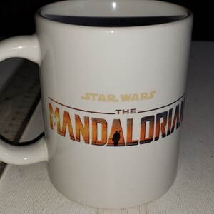 May include: White ceramic mug featuring a black and gold graphic of the Star Wars: The Mandalorian logo. The logo depicts a sunset scene with a silhouette of a person in the centre.