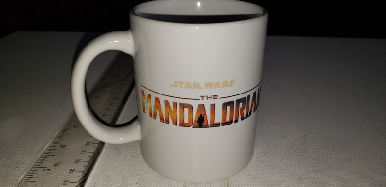 May include: White ceramic mug featuring a black and gold graphic of the Star Wars: The Mandalorian logo. The logo depicts a sunset scene with a silhouette of a person in the centre.