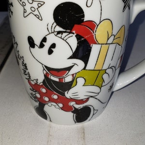 May include: White ceramic mug with a black and red illustration of Minnie Mouse holding a yellow and green gift box. The mug has a black rim and a black handle.