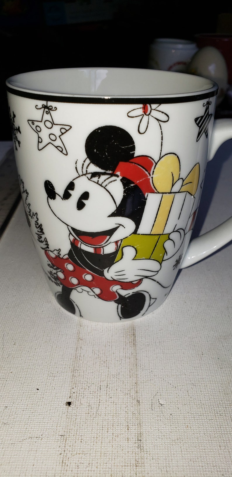 May include: White ceramic mug with a black and red illustration of Minnie Mouse holding a yellow and green gift box. The mug has a black rim and a black handle.
