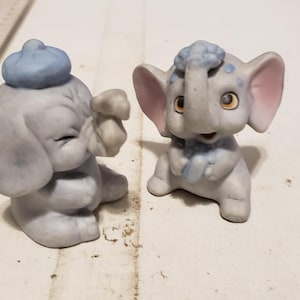 May include: Two small ceramic elephant figurines. One elephant is wearing a blue hat and the other has blue and pink accents.