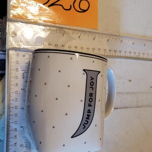 K&ouml;nnte beinhalten: Wei&szlig;e Keramik-Tasse mit schwarzem Rand und Henkel. Die Tasse hat einen grauen Buchstaben "J" mit den Worten "JUMP FOR JOY" und kleinen schwarzen Punkten. Ein Lineal befindet sich neben der Tasse.