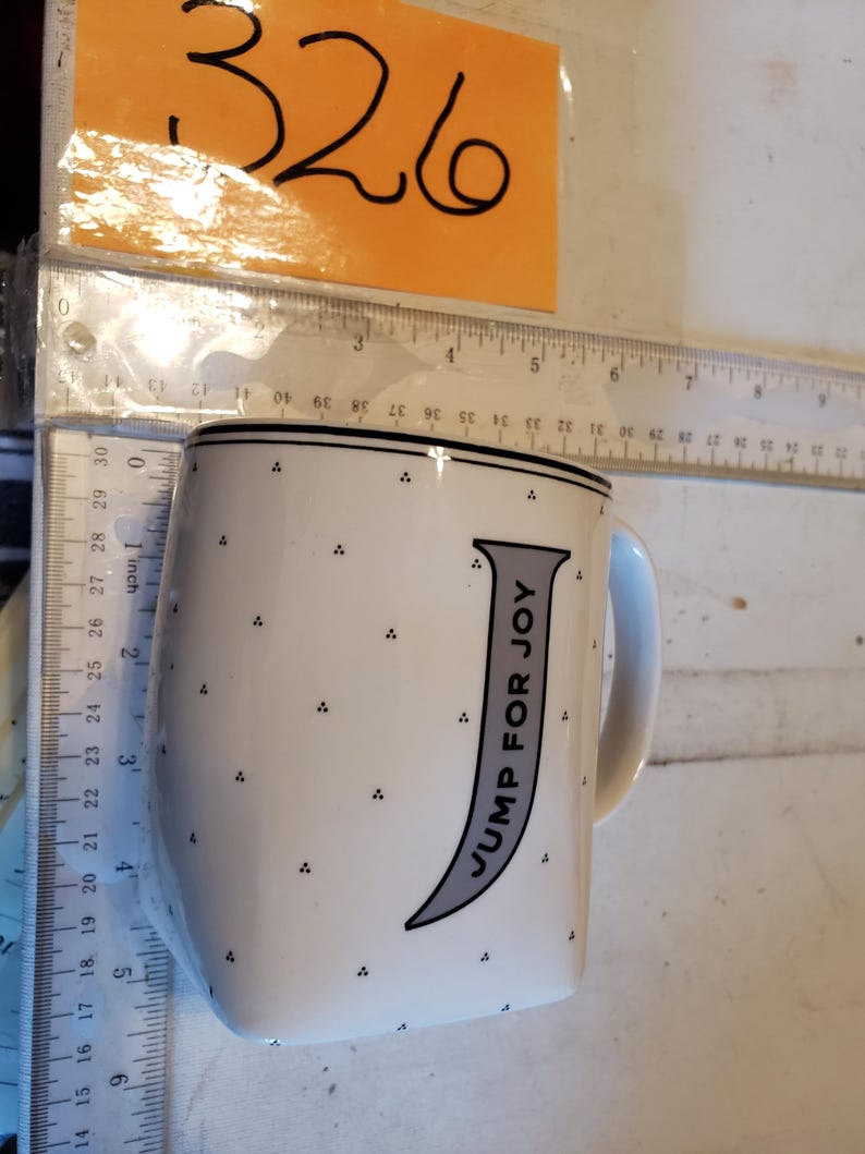K&ouml;nnte beinhalten: Wei&szlig;e Keramik-Tasse mit schwarzem Rand und Henkel. Die Tasse hat einen grauen Buchstaben "J" mit den Worten "JUMP FOR JOY" und kleinen schwarzen Punkten. Ein Lineal befindet sich neben der Tasse.