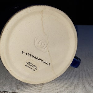 Pu&ograve; includere: Il fondo di una tazza in ceramica blu con la scritta "by Anthropologie" e "Made in China" stampata sul fondo.