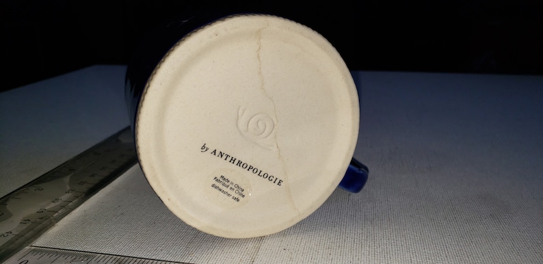 Pu&ograve; includere: Il fondo di una tazza in ceramica blu con la scritta "by Anthropologie" e "Made in China" stampata sul fondo.