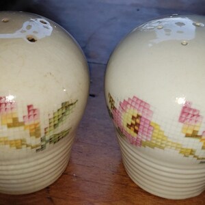 May include: A pair of white ceramic salt and pepper shakers with a floral design. The shakers have a round top with holes for dispensing salt and pepper. The floral design features pink and yellow flowers with green leaves.