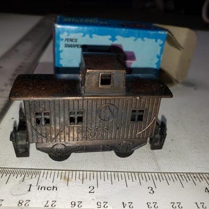 May include: A vintage metal pencil sharpener shaped like a train caboose. The caboose is bronze-colored with etched details, including windows and the text "S & S 1346". A blue box with "PENCIL SHARPENER" is in the background.