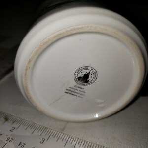 以下が含まれることがあります： 白いセラミック製のマグカップの底面。黒いディズニーのロゴがあり、「Authentic Original Disney Theme Parks」と「Disney 中国製 食洗機対応 電子レンジ対応」と書いてあります。