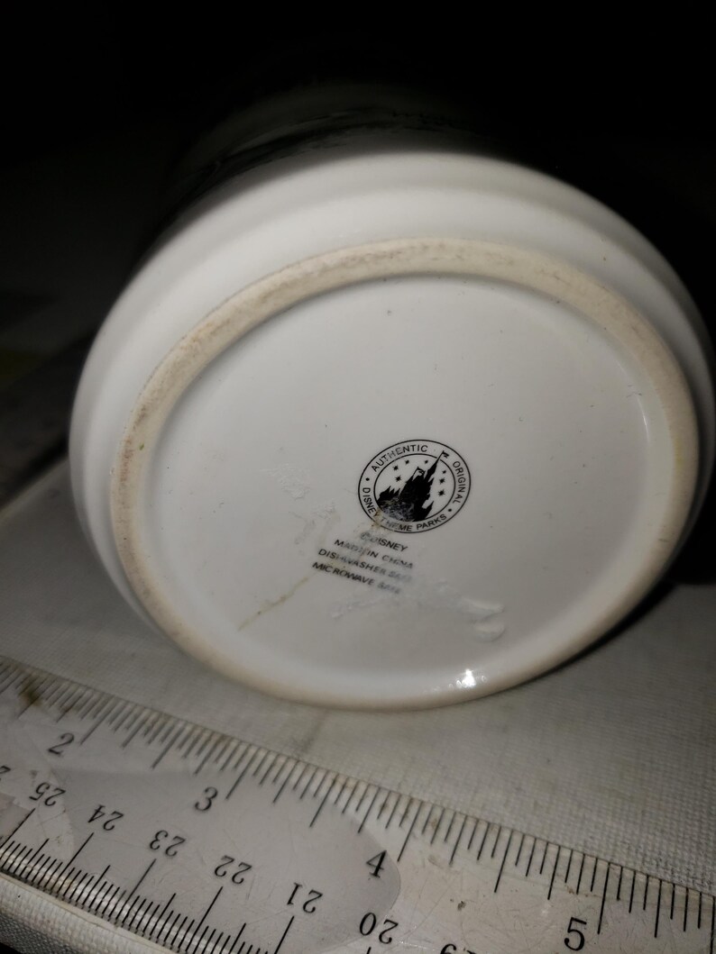 以下が含まれることがあります： 白いセラミック製のマグカップの底面。黒いディズニーのロゴがあり、「Authentic Original Disney Theme Parks」と「Disney 中国製 食洗機対応 電子レンジ対応」と書いてあります。