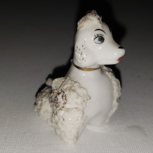 May include: White porcelain figurine of a poodle with gold trim around the neck. The poodle is sitting with its head turned to the side and has painted eyes and red lips.