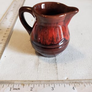 May include: A small, brown ceramic creamer with a handle and spout. The creamer has a glossy finish with a reddish-brown color gradient. A ruler with inch measurements is in the foreground.