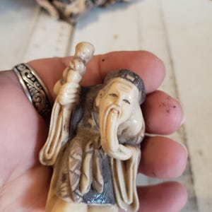 May include: A small, intricately carved figurine of an elderly man with a long beard and a staff. The figure is primarily cream-colored with accents of gray and brown, and is held in a hand.