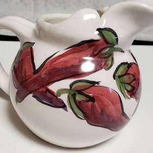 May include: White ceramic pitcher with a red and green floral design. The pitcher has a rounded body and a handle.