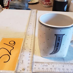 K&ouml;nnte beinhalten: Wei&szlig;er Keramikbecher mit gebogenem Henkel, mit dem Text "JUMP FOR JOY" in Blau. Der Becher hat einen schwarzen Rand und kleine graue Dreieckmuster. Ein Lineal zeigt cm-Messungen.