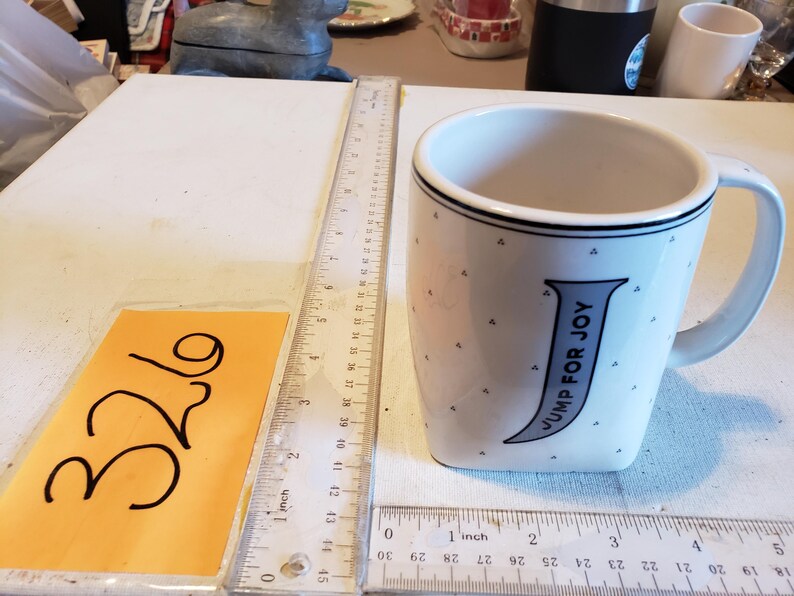 K&ouml;nnte beinhalten: Wei&szlig;er Keramikbecher mit gebogenem Henkel, mit dem Text "JUMP FOR JOY" in Blau. Der Becher hat einen schwarzen Rand und kleine graue Dreieckmuster. Ein Lineal zeigt cm-Messungen.