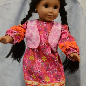 May include: A doll wearing a pink dress with a floral pattern and peace signs. The dress has a pink vest with a tie-dye pattern. The doll has long black braids and is wearing earrings.