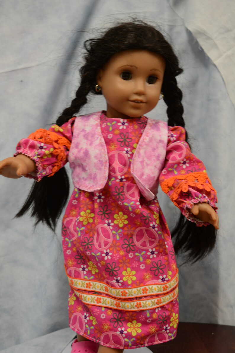 May include: A doll wearing a pink dress with a floral pattern and peace signs. The dress has a pink vest with a tie-dye pattern. The doll has long black braids and is wearing earrings.