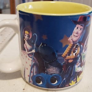 May include: A white ceramic mug with a blue background and a yellow rim. The mug features a colorful image of characters from the Toy Story movie, including Woody, Buzz Lightyear, and Jessie.