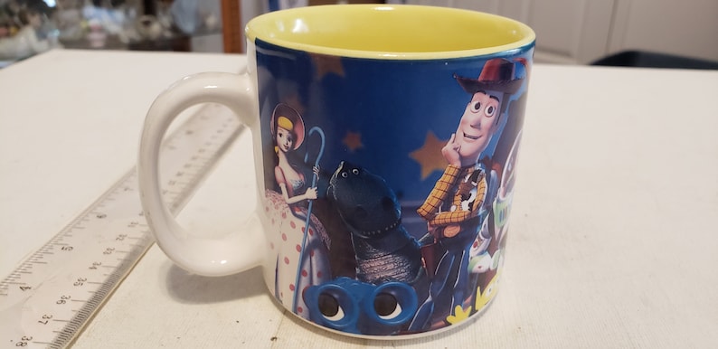 May include: A white ceramic mug with a blue background and a yellow rim. The mug features a colorful image of characters from the Toy Story movie, including Woody, Buzz Lightyear, and Jessie.