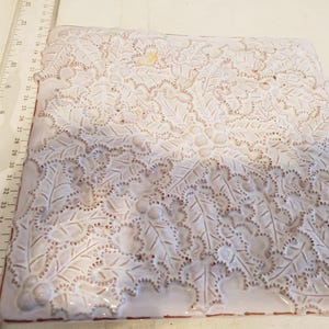 May include: A white ceramic tile with a raised design of holly leaves and berries. The tile has a slightly textured surface and a light brown border.
