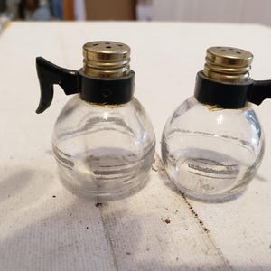 May include: Two clear glass salt and pepper shakers with black plastic handles and gold colored tops.
