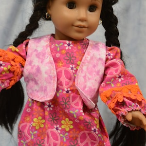 May include: A doll wearing a pink and white vest over a pink dress with a peace sign and floral pattern. The doll has long black hair in braids and brown eyes.