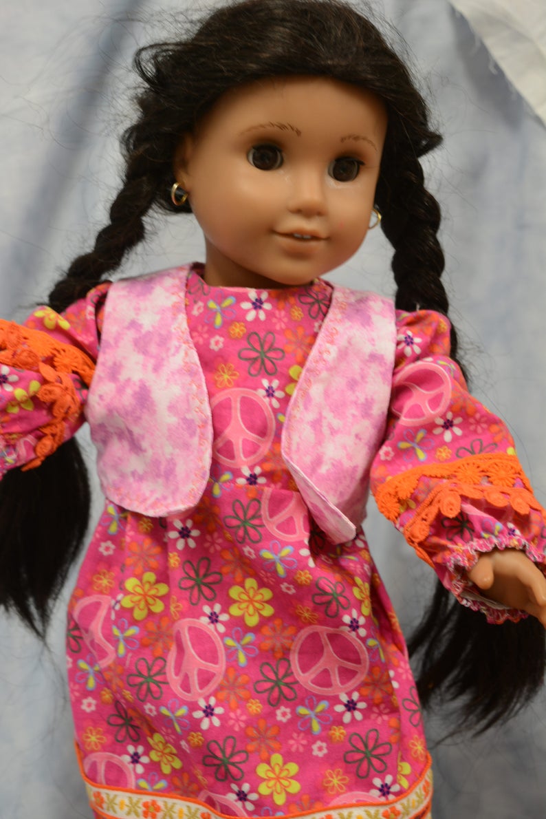 May include: A doll wearing a pink and white vest over a pink dress with a peace sign and floral pattern. The doll has long black hair in braids and brown eyes.