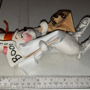 Puede incluir: Un fantasma de peluche blanco con un sombrero rojo y naranja, sosteniendo una bandera blanca con la palabra "BOO" escrita en ella. El fantasma lleva una bolsa de papel marr&oacute;n en la cabeza y tiene un palo en la mano.