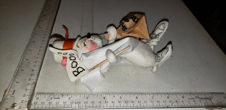 Puede incluir: Un fantasma de peluche blanco con un sombrero rojo y naranja, sosteniendo una bandera blanca con la palabra "BOO" escrita en ella. El fantasma lleva una bolsa de papel marr&oacute;n en la cabeza y tiene un palo en la mano.