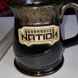 May include: Black ceramic mug with a brown speckled glaze. The mug has a handle and a rectangular label that reads "Boondock Nation" with a mountain range graphic.