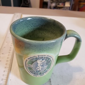 May include: Green ceramic mug with a handle. The mug has a two-tone color scheme, with a darker green rim and a lighter green body. A circular emblem on the front reads "The Original Pancake House" and "Since 1953".