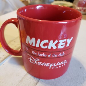 Puede incluir: Una taza de cer&aacute;mica roja con un asa curva. La taza presenta la palabra "MICKEY" en letras blancas, junto con las frases "the leader of the club" y "Disneyland PARIS" en escritura blanca. La taza es de color rojo liso.