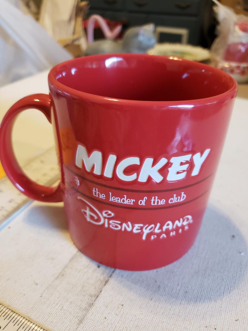 Puede incluir: Una taza de cer&aacute;mica roja con un asa curva. La taza presenta la palabra "MICKEY" en letras blancas, junto con las frases "the leader of the club" y "Disneyland PARIS" en escritura blanca. La taza es de color rojo liso.