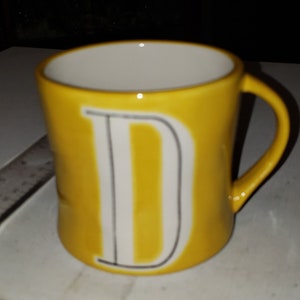 Op de afbeelding: Een gele keramische mok met een witte letter "D" aan de voorkant.