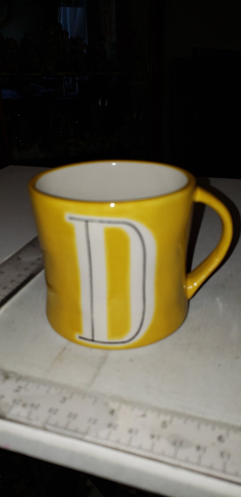 Op de afbeelding: Een gele keramische mok met een witte letter "D" aan de voorkant.