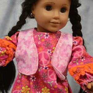May include: A doll with long black braids wearing a pink and white vest over a pink dress with peace signs and flowers. The doll has brown eyes and is wearing gold hoop earrings.