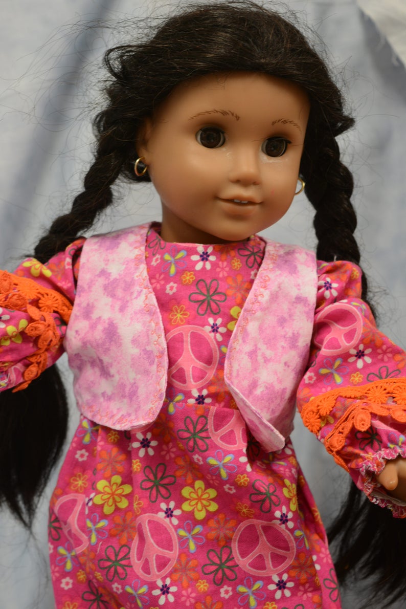 May include: A doll with long black braids wearing a pink and white vest over a pink dress with peace signs and flowers. The doll has brown eyes and is wearing gold hoop earrings.