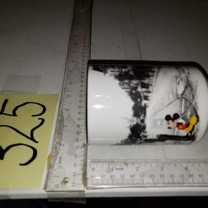 以下が含まれることがあります： 白地に黒と白のミッキーマウスとミニーマウスの冬景色が描かれた白いセラミックマグカップ。マグカップは黒の縁取りと取っ手です。