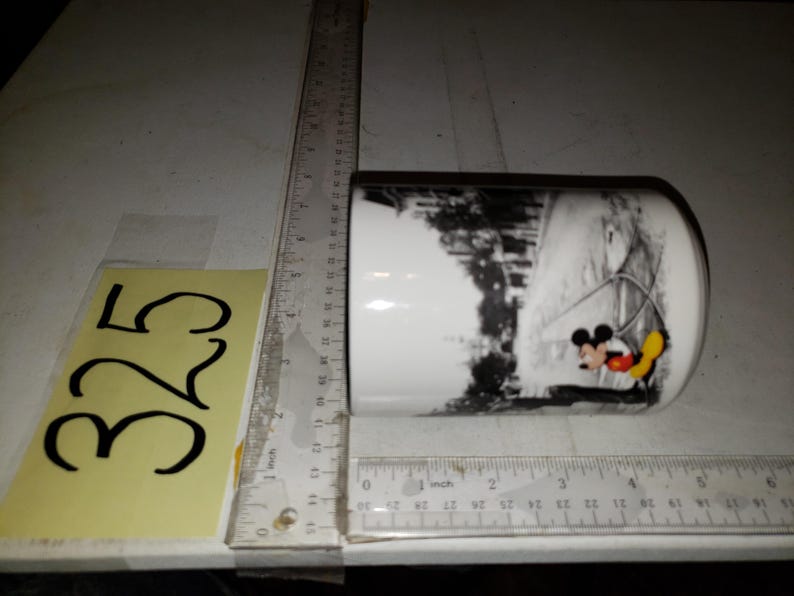 以下が含まれることがあります： 白地に黒と白のミッキーマウスとミニーマウスの冬景色が描かれた白いセラミックマグカップ。マグカップは黒の縁取りと取っ手です。