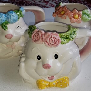 May include: Three white ceramic mugs with animal faces and floral designs. Each mug has a different floral design with pink, blue, and yellow flowers. The mugs have handles and are perfect for a tea party or a special occasion.