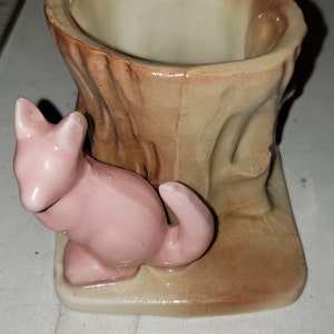 May include: A ceramic planter shaped like a tree stump with a pink squirrel figurine sitting on the base. The planter has a light brown glaze with a darker brown glaze on the tree bark texture.