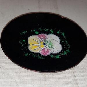 May include: A small black plate with a painted flower design. The flower is pink, yellow, and white with green accents.
