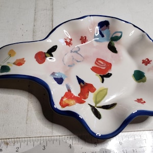 May include: White ceramic spoon rest with a blue rim and a floral design. The spoon rest features red, yellow, and blue flowers with green leaves. The spoon rest is approximately 8 inches long.