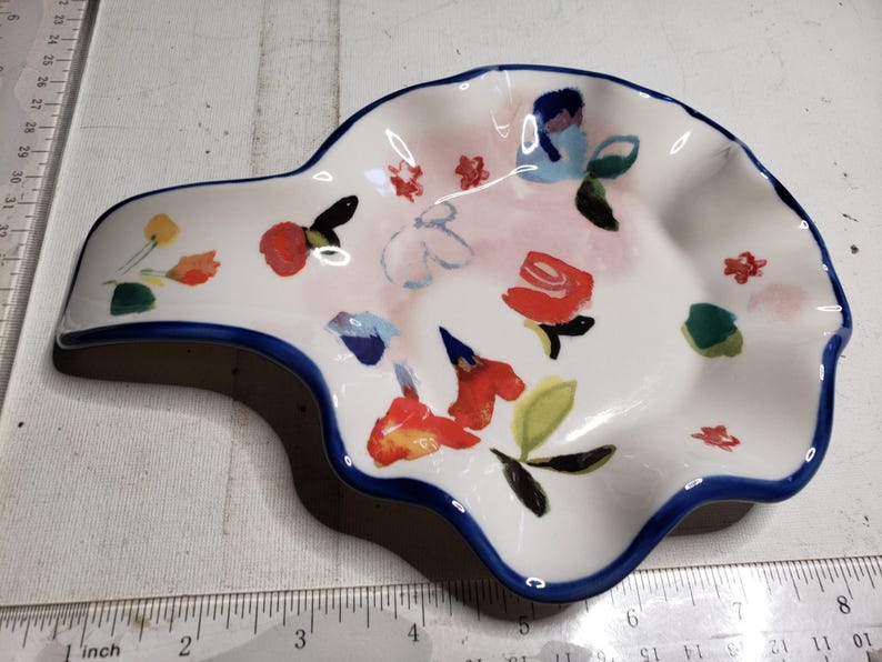 May include: White ceramic spoon rest with a blue rim and a floral design. The spoon rest features red, yellow, and blue flowers with green leaves. The spoon rest is approximately 8 inches long.